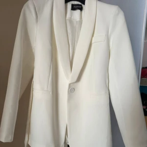 Vit kavaj med bälte - Stilren vit kavaj  med klassisk krage, knäppning framtill och tillhörande bälte i midjan. Kavajen har raka ärmar, två fickor och en snygg, minimalistisk look. Perfekt för dig som gillar en clean och modern stil. Helt ny och oanvänd. Passar perfekt att ha till studenten eller andra festliga tillfällen.