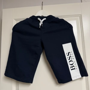 Marinblå shorts från Hugo Boss - Snygga marinblå shorts från Hugo Boss i mjuk bomull med vit snörning i midjan och stor vit logga på ena benet. Perfekta för chill dagar eller när du vill ha en clean och sportig look.