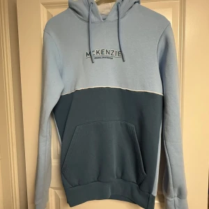 Ljusblå hoodie från McKenzie - Snygg hoodie från McKenzie i två nyanser av blått. Tröjan har huva med snörning, stor magficka och broderad logga på bröstet. Perfekt för chill dagar och enkel att matcha med jeans eller joggers.