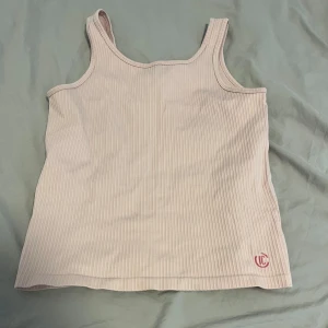Ljusbeige ribbad linnetopp LC - Säljer en ljusbeige ribbad linnetopp från LC med broderad logga i rött nertill. Toppen har breda axelband och ärmlös design, perfekt för varma dagar. Enkel och stilren med en mjuk känsla och stretchigt material.