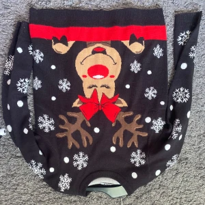 Svart jultröja med renmotiv och snöflingor - Svart stickad jultröja med ett stort motiv av en ren med röd rosett och röd mule framtill. Tröjan har vita snöflingor och prickar över hela, samt långa ärmar. Perfekt för att maxa julkänslan med ett lekfullt och färgglatt print. Andvänd 2 gånger. Pris kan diskuteras.