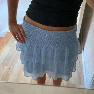 Ljusblå volangkjol med blommor - Supersöt ljusblå kortkjol med volanger och smock i midjan. Kjolen är gjord i ett tunt, flowy material som känns perfekt för sommaren. Passar dig som gillar en feminin och trendig stil.