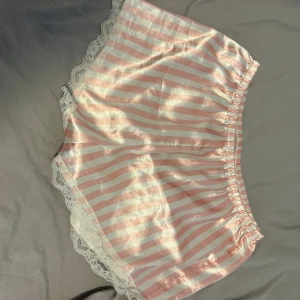 Rosa och vita randiga satinsleep-shorts - Supergulliga shorts i glansig satin med rosa och vita ränder. De har elastisk midja och är dekorerade med vit spets längs kanterna för en extra söt vibe. Perfekta för mysiga hemmakvällar eller som pyjamasshorts.