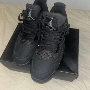 Svarta Nike Air Jordan 4 sneakers - Säljer ett par helsvarta Nike Air Jordan 4 sneakers med klassisk Jumpman-logga på plös och häl. Skorna har snörning, meshdetaljer och robust sula med vit logga undertill. Ovandelen är i mocka och syntet, vilket ger en clean och sportig look.