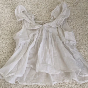 Vit volangblus från Gina Tricot - Supersöt vit blus från Gina Tricot i luftig bomull. Toppen har volangdetaljer vid axlarna, knytning i ryggen och en flowy, lätt A-linjeform. Perfekt för en somrig look med romantiska vibes.