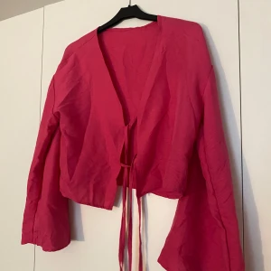 Rosa omlottopp med knytband - Snygg rosa omlottopp med långa ärmar och croppad passform. Toppen har knytband framtill som ger en trendig look och är tillverkad i ett lätt, luftigt material. 