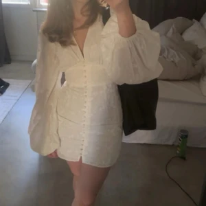 Vit kort klänning med puffärmar - Superfin vit kort klänning med v-ringning, markerad midja och stora puffärmar. Klänningen har knappar framtill och ett diskret blommigt mönster i tyget. Från adoore och endast använd 1 gång, nypris 1400