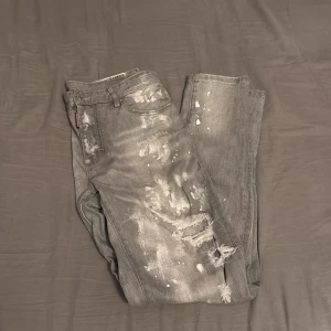 Grå Dsquared2 jeans med slitningar - Säljer ett par grå jeans från Dsquared2 med coola slitningar och färgstänk för en edgy look. Jeansen har klassisk femficksdesign, dragkedja och patch med Dsquared2-logga bak. Perfekta för dig som gillar streetstyle och vill sticka ut.