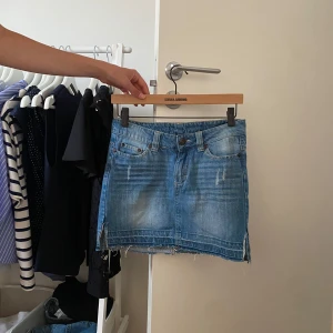 Blå jeanskjol med slitna detaljer - Snygg blå jeanskjol med slitna detaljer och rå kant nertill. Klassisk femficksmodell med knapp och dragkedja framtill. Slits på sidorna. Perfekt för dig som gillar en avslappnad och trendig stil.