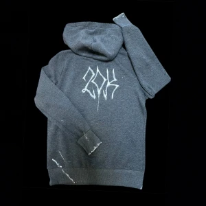 Project 20K Hoodie - Jättefin custom made mörkgrå hoodie med vit text på ryggen, slitningar samt färgstänk på ärmar och framsida. Den har en klassisk huva  och känguruficka framtill. Perfekt för dig som är intresserad av fashion och unika detaljer.