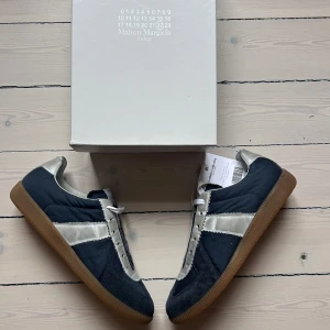 Maison Margiela sneakers i blått och silver - Maison Margiela sneakers med marinblå mocka och textil, silverfärgade detaljer i skinn och klassisk gummisula. Snygg kontrast mellan färgerna och ikonisk design med vita snören och märkets logga på plösen. Perfekta för dig som gillar stilrena men unika sneakers. Tillkommer Box och dustbag + authentication bevis från Vestiaire Collective. (HELT NYA)