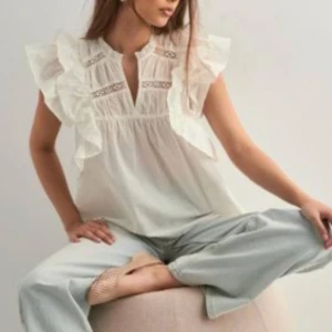 Vit blus med volang och spetsdetaljer - Superfin vit blus med volangiga ärmar och snygga spetsdetaljer vid axlar och bröst. Luftig modell med v-ringning och lätt transparent tyg, perfekt för en fräsch och trendig look. Bara testad.