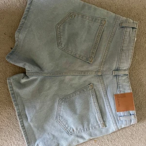 Ljusblå jeansshorts från Never Denim - Säljer ett par ljusblå jeansshorts från MH med hög midja. Shortsen har bälteshällor, knapp och dragkedja framtill samt en snygg läderpatch bak. Perfekta för sommaren och riktigt sköna att bära. Midjemåttet är 35 och är storlek s