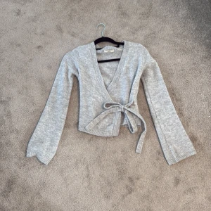 Grå omlottkofta från Gina Tricot - Superfin ljusgrå omlottkofta i storlek xs från Gina Tricot med knyt i midjan och v-ringning. Koftan är stickad, har långa lösare ärmar och en croppad passform som ger en trendig look. Perfekt att slänga över en topp för något extra! Använd fåtal gånger så i nästintill nyskick💓Pris går att diskutera!💖