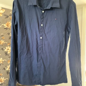 Mörkblå skjorta från Tommy Hilfiger - Klassisk mörkblå skjorta från Tommy Hilfiger med vit knappstängning och diskret broderad logga på bröstet. Skjortan har lång ärm, normal passform och är tillverkad i mjuk bomull. Perfekt för dig som gillar stilrena och tidlösa plagg. Tveka inte att fråga om du vill ha fler bilder eller några frågor ! Priset är ej hugget i sten 