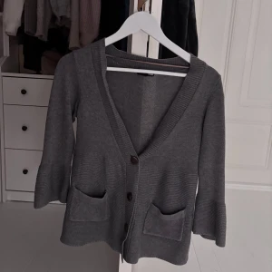 Grå kofta med fickor från H&M - Mysig grå kofta från H&M med trekvartsärmar, djup v-ringning och knappar framtill. Koftan har två praktiska fickor och är stickad i mjukt material. Perfekt att slänga över en t-shirt eller linne för en chill look.