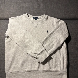 Grå sweatshirt från Polo Ralph Lauren - Klassisk grå sweatshirt från Polo Ralph Lauren med rund halsringning och broderad marinblå logga på bröstet. Tröjan har långa ärmar och ribbade muddar vid ärmslut och nederkant. Tröjan har endast små röda fläckar som kan tas bort med kemtvätt, storlek 152