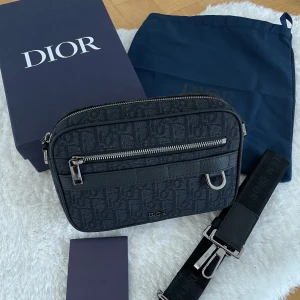 Svart axelväska från Dior med monogram - Snygg svart axelväska från Dior med klassiskt monogrammönster och silverfärgade detaljer. Väskan har justerbar axelrem, dragkedja framtill och på toppen samt en stilren, rektangulär form. Perfekt för dig som vill ha en exklusiv och trendig accessoar.
