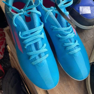 Blå adidas fotbollsskor strl 38 - Säljer ett par snygga blå adidas fotbollsskor med rosa detaljer och vita dobbar. Skorna har snörning och är gjorda i syntetmaterial med en smal, sportig siluett. Perfekta för dig som vill sticka ut på planen med stil.