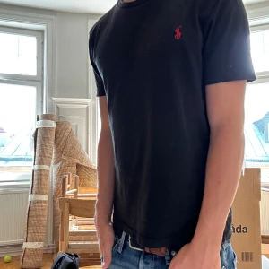 Svart t-shirt från Polo Ralph Lauren - Svart t-shirt från Polo Ralph Lauren med röd broderad logga på bröstet. Modellen är slim fit och har klassisk rund hals och korta ärmar. Tillverkad i mjuk bomull som känns skön mot huden. Perfekt för en clean och stilren look.
