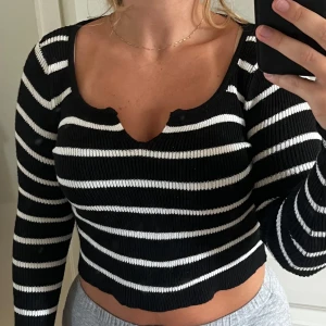 Svartvit randig croppad tröja - Säljer en svart och vit randig croppad tröja med lång ärm och ribbad struktur. Tröjan har en snygg v-ringning med liten cutout-detalj framtill och är perfekt för dig som gillar en trendig och bekväm stil.