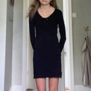 Svart kortklänning från Tessie - Supersnygg svart kortklänning från Tessie med lång ärm och snörning framtill vid halsringningen. Klänningen har en tight passform och är perfekt för dig som gillar en stilren look. Materialet känns mjukt och stretchigt, vilket gör den bekväm att bära.