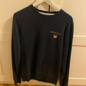Mörkblå sweatshirt från GANT - Snygg mörkblå sweatshirt från GANT med broderad logga och sköld på bröstet. Klassisk rund halsringning och långa ärmar. Perfekt för dig som gillar stilrena och tidlösa plagg. Tillverkad i mjuk bomull för skön komfort.