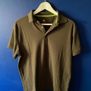 Olivgrön pikétröja från McKinley - Snygg olivgrön pikétröja från McKinley i klassisk modell med krage och två knappar. Tröjan har korta ärmar och en diskret grön detalj på insidan av kragen. Perfekt för en avslappnad och sportig stil.