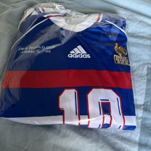 Frankrike VM 98 Adidas fotbollströja - Klassisk blå fotbollströja från Adidas, officiell Frankrike FIFA World Cup 1998. Tröjan har vita och röda detaljer, breda ränder och nummer 10 på framsidan. Broderad tupp-logga och text på bröstet. Perfekt för dig som älskar retro och fotbollshistoria.