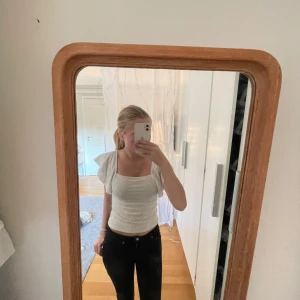 Vit broderad blus med volangärm - Superfin vit blus med fyrkantig ringning och korta volangärmar. Toppen har broderade detaljer och är gjord i ett lätt bomullstyg som känns luftigt. Perfekt att styla med jeans eller kjol för en fräsch look.