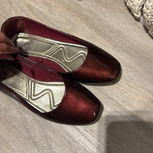 Vinröda ballerinaskor - Snygga ballerinaskor i vinröd metallic med fyrkantig tå och låg platt sula. Skorna har en glansig finish och insida i kontrasterande guldton. Perfekta för dig som vill sticka ut med en elegant men ändå chill look.