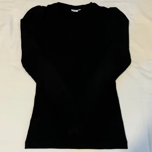 Svart långärmad tröja - Svart ribbad långärmad tröja med puffade axlar och rund halsringning. Tröjan är figurnära och har en stilren look som passar perfekt till jeans eller kjol. Mjuk och stretchig i materialet för extra komfort.