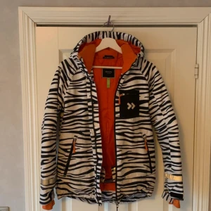 Zebramönstrad skid jacka från 157 Funk - Cool zebramönstrad skidjacka från 157 Funk med svartvita ränder och orange foder. Jackan har huva, dragkedjor och reflex på ärmarna. Perfekt för dig som i vinter vill hålla dig varm med stil.