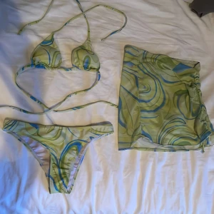 Grön och blå swirl bikini med kjol - Trendigt bikini-set med swirlmönster i grönt, blått och gult. Triangeltopp med knyt och matchande bikiniunderdel. Ingår även en meshkjol med dragsko i samma mönster som coverup. Perfekt för stranden och poolhäng.