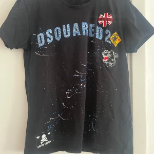 Svart Dsquared2 t-shirt med tryck - Cool svart t-shirt från Dsquared2 med blått logotryck, patchar och färgstänk framtill. Unik design med pantermärke, brittisk flagga och Caten Twins-detalj. Skön passform och tillverkad i mjuk bomull. Perfekt för dig som gillar streetstyle och statement-plagg.