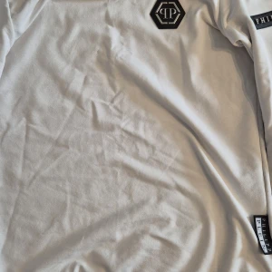 Vit hoodie från Philipp Plein - Vit hooide från Philipp Plein med svart logotypmärke på bröstet och ett svart band med märkesnamnet på ärmen. Klassisk passform och mjukt bomullsmaterial, perfekt för en clean och stilren look. Den är storlek M men passar jättebra S