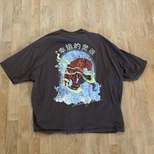 Brun oversized t-shirt med tigertryck - Cool brun t-shirt från ASOS DESIGN med stort tryck av en tiger och asiatiska tecken på ryggen. Oversized fit och klassisk rund hals. Tillverkad i mjuk bomull. 