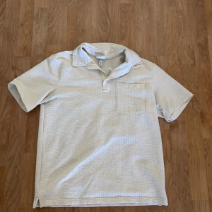 Vit pikétröja från H&M i bomull - Säljer en vit pikétröja från H&M i regular fit. Tröjan har korta ärmar, klassisk krage och en bröstficka. Materialet är bomull med en diskret strukturerad yta. 