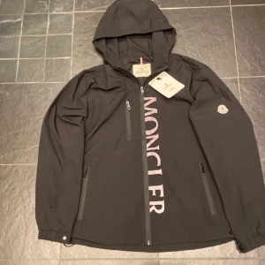 Svart vindjacka från Moncler med huva - Svart vindjacka från Moncler med stor logga framtill och klassisk patch på ärmen. Jackan har huva, dragkedjor på fickor och bröst, samt snygga detaljer som tryckknappar vid ärmslut. Perfekt för dig som vill ha en clean och sportig look.