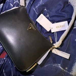 Svart axelväska från Guess - Snygg svart axelväska från Guess med klassisk trekantslogga i metall framtill. Väskan har guldfärgade detaljer, dragkedja upptill och ljusbeige foder med logomönster. Perfekt storlek för det viktigaste och stilren design som passar till allt.