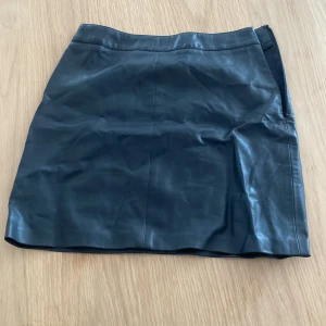 Svart skinnkjol från Gina Tricot - Snygg svart kort skinnkjol från Gina Tricot i storlek 34. Kjolen har en rak passform och dragkedja i sidan för enkel påtagning. 