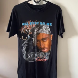 Svart 2Pac t-shirt med tryck - Säljer en svart t-shirt med stort färgglatt tryck av 2Pac och texten 'All Eyez On Me' på bröstet. T-shirten har klassisk passform, rund hals och korta ärmar. Perfekt för dig som gillar hiphop och streetwear.
