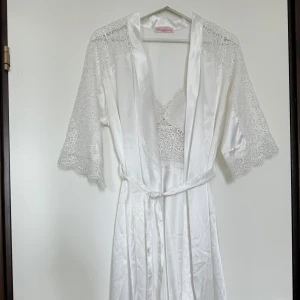 Vit spetsklänning i satin från Mariposa - Superfin vit lång klänning i satin med tunna axelband och vacker spets upptill. Tillhörande kimono med spetsdetaljer på ärmarna och knytband i midjan. Perfekt för dig som vill ha en romantisk och lyxig look.