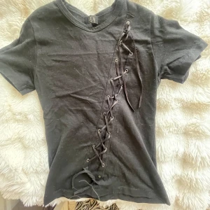 Svart t-shirt med snörning och tryck - Svart t-shirt från J.W. med snörningsdetalj diagonalt framtill och två röda draktryck på ryggen. T-shirten har rund halsringning och korta ärmar. Materialet känns som bomull och passformen är normal. Grym statement-piece för dig som gillar edgy stil. Köpt i Tokyo🥰