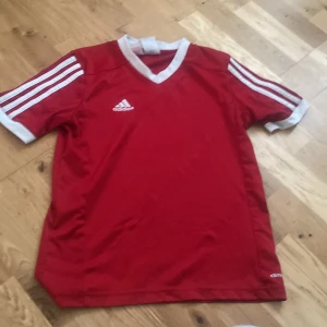 Röd Adidas fotbollströja med vita detaljer - Klassisk röd fotbollströja från Adidas med vita ränder på ärmarna och vit V-ringad krage. Tröjan har broderad Adidas-logga på bröstet och är tillverkad i lätt och ventilerande material, perfekt för träning eller match. Ligger i Vetlanda 