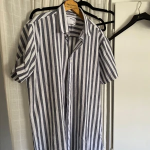 Linneskjorta Herr - HELT NY! - Snygg kortärmad linneskjorta från Selected med breda vertikala ränder i vitt och ljusblått. Den är använd 2 gånger bara då jag köpte många nya skjortor! Skjortan har klassisk krage och knäppning framtill. Nypris är 499kr, men jag säljer den för endast 299! Jag kan gå ner i pris vid snabb affär, hör gärna av dig.