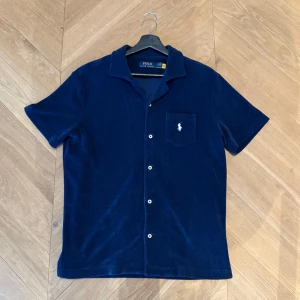 Ralph Lauren tröja - Mörkblå skjorta från Polo Ralph Lauren i mjuk frotté, kortärmad med klassisk krage och knappar framtill. Bröstficka med broderad vit logga ger en clean och sportig vibe. Perfekt för dig som gillar stilren och avslappnad look. Pris kan diskuteras