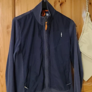 Blå vindjacka från Parajumpers - Snygg blå vindjacka från Parajumpers med dragkedja framtill och ribbad krage. Jackan har en cool patch på ärmen och orange detaljer vid halsen. Perfekt för dig som vill ha en stilren och sportig look. Fickor med dragkedja och smidig passform.