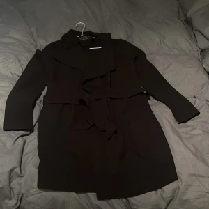Svart kappa från ONLY med bälte - Svart kappa från ONLY med snygg omlottkrage och bälte i midjan. Jackan har lång ärm och en stilren, klassisk look som passar perfekt till lager-på-lager. Materialet känns mjukt och följsamt, och designen är enkel men trendig. Litet hål som jag tagit bild på men syns inte om man inte tittar på nära håll. 