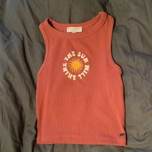 Rostfärgad ribbad linnetopp XS - Snygg rostfärgad ribbad linnetopp från Pacific Republic. Framtill finns ett broderat gult solmotiv och texten 'The Sun Will Shine' i vitt. Toppen är ärmlös och har en tight passform, perfekt för varma dagar.
